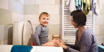 Un enfant est sur les toilettes d’une salle de bain face à sa mère qui lui apprend la propreté.