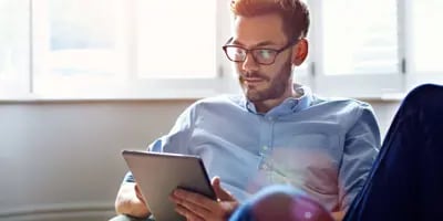 Un homme brun avec des lunettes regarde sa tablette. Derrière lui se trouve un ensemble de fenêtres blanches.