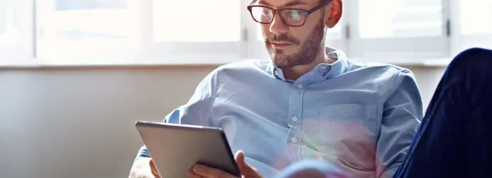 Un homme brun avec des lunettes regarde sa tablette. Derrière lui se trouve un ensemble de fenêtres blanches.