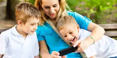 Une mère et ses deux enfants souriants sont assis sur un banc en plein air et regardent ensemble un écran de téléphone.
