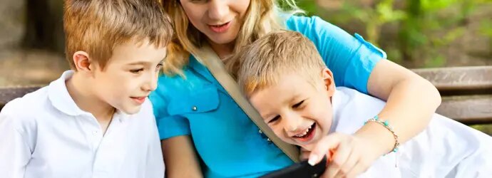 Une mère et ses deux enfants souriants sont assis sur un banc en plein air et regardent ensemble un écran de téléphone.