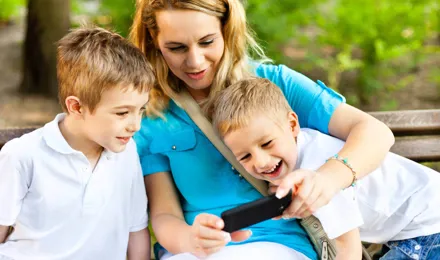Une mère et ses deux enfants souriants sont assis sur un banc en plein air et regardent ensemble un écran de téléphone.