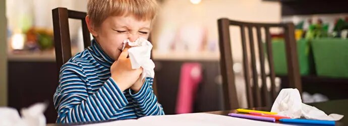 Un enfant a le nez qui coule et se mouche. Il est assis devant un cahier, des crayons de couleur et un mouchoir usagé. 
