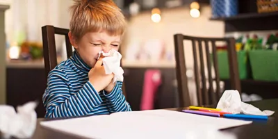 Un enfant a le nez qui coule et se mouche. Il est assis devant un cahier, des crayons de couleur et un mouchoir usagé. 
