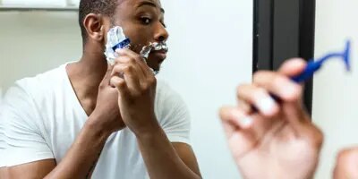 Un homme est en train de se raser avec une mousse à raser étalée sur sa barbe. Il se regarde dans le miroir.  