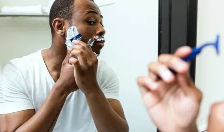 Un homme est en train de se raser avec une mousse à raser étalée sur sa barbe. Il se regarde dans le miroir.  