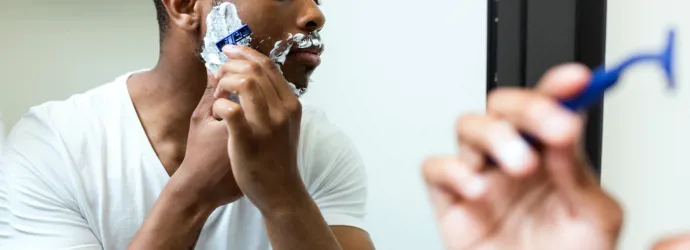 Un homme est en train de se raser avec une mousse à raser étalée sur sa barbe. Il se regarde dans le miroir.  