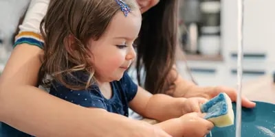 Un enfant apprend à faire la vaisselle avec l'aide d'un adulte souriant.