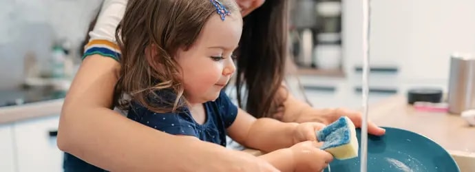 Un enfant apprend à faire la vaisselle avec l'aide d'un adulte souriant.