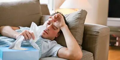Un homme malade est couché sur un canapé beige. Il a la main posée sur son front et une boîte de mouchoirs sur lui.  