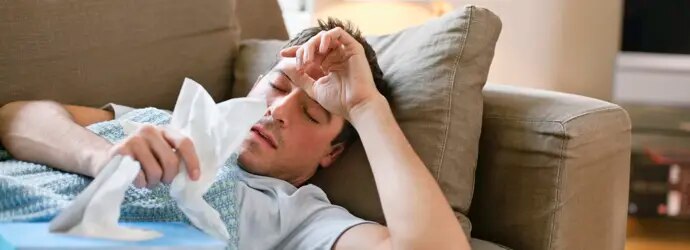Un homme malade est couché sur un canapé beige. Il a la main posée sur son front et une boîte de mouchoirs sur lui.  
