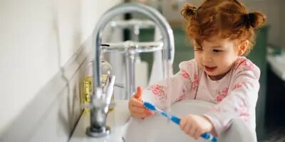 Une petite fille rousse est au bord d’un lavabo avec une brosse à dents dans la main qu’elle regarde. 