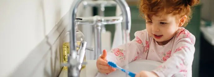 Une petite fille rousse est au bord d’un lavabo avec une brosse à dents dans la main qu’elle regarde. 
