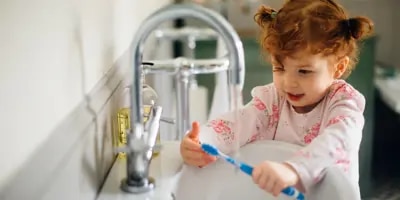 Une petite fille rousse est au bord d’un lavabo avec une brosse à dents dans la main qu’elle regarde.