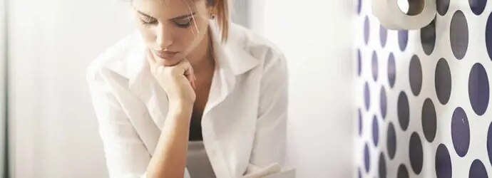 Une femme réfléchit assise aux toilettes à côté d’un mur à pois et d’un rouleau de papier toilette. 
