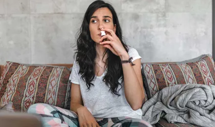 Koivun siitepölyallergiasta kärsivä nuori nainen istuu sohvalla pidellen nenäsumutetta nenäänsä vasten
