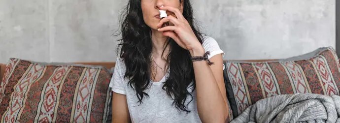 Une jeune femme avec une allergie au pollen de bouleau est assise sur le canapé et porte un spray nasal à son nez