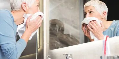 Une femme avec des  cheveux blancs et courts regarde dans le miroir de salle de bain après avoir rincé son visage pour soulager ses yeux larmoyants à cause de l'allergies