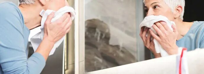 Une femme avec des  cheveux blancs et courts regarde dans le miroir de salle de bain après avoir rincé son visage pour soulager ses yeux larmoyants à cause de l'allergies