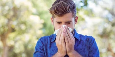 Un jeune homme dans une chemise bleue avec un fond de forêt floue derrière lui éternue et a les symptômes du rhume des foins