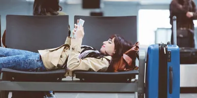Une femme installée sur plusieurs sièges dans un aéroport lit un livre avec une valise à roulettes à côté d'elle