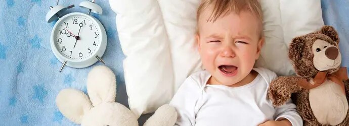 Un bébé pleure sur une couverture entourée de deux jouets en peluche et d'un réveil analogique