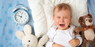 Un bébé pleure sur une couverture entourée de deux jouets en peluche et d'un réveil analogique