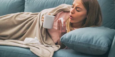 Une femme est allongée sur son canapé et souffre de toux persistante. Elle tient un mouchoir dans sa main et une tasse.