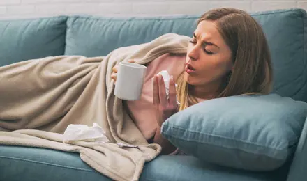 Une femme est allongée sur son canapé et souffre de toux persistante. Elle tient un mouchoir dans sa main et une tasse.