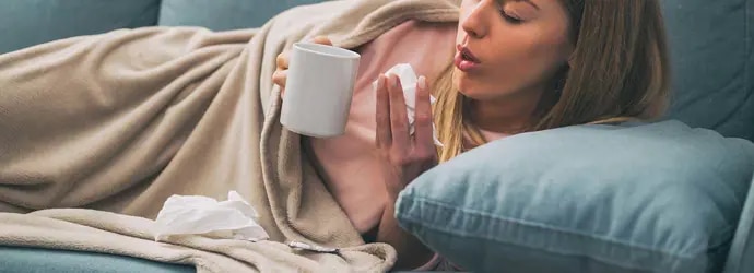 Une femme est allongée sur son canapé et souffre de toux persistante. Elle tient un mouchoir dans sa main et une tasse.