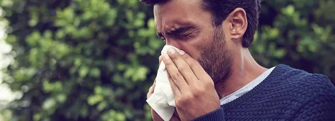 Un homme se mouche avec un mouchoir tout en ayant les  yeux larmoyants et le nez qui coule