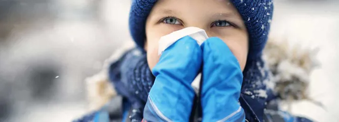 Photo d’un enfant qui se mouche. Il est dehors sous la neige et porte un bonnet et des moufles.