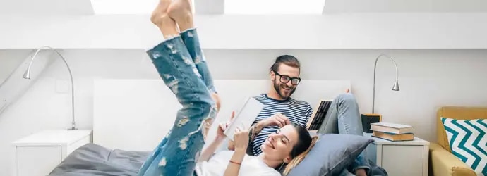 Un jeune couple assis sur un lit, lisent dans une chambre avec deux puits de lumière