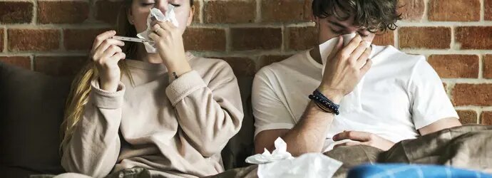 Deux jeunes adultes sont assis sous des couvertures et éternuent dans des mouchoirs avec un mur de briques derrière eux