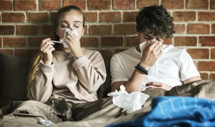 Deux jeunes adultes sont assis sous des couvertures et éternuent dans des mouchoirs avec un mur de briques derrière eux