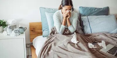 Une femme est dans son lit et se mouche entourée de mouchoirs utilisés, souffrant probablement d’un rhume interminable.