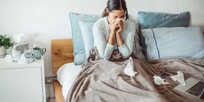 Une femme est dans son lit et se mouche entourée de mouchoirs utilisés, souffrant probablement d’un rhume interminable.