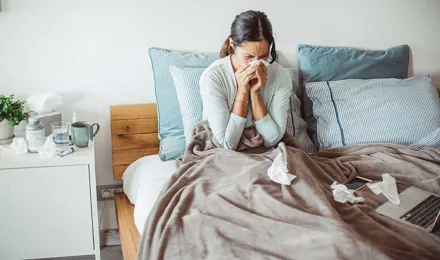 Une femme est dans son lit et se mouche entourée de mouchoirs utilisés, souffrant probablement d’un rhume interminable.