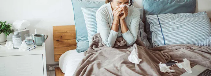 Une femme est dans son lit et se mouche entourée de mouchoirs utilisés, souffrant probablement d’un rhume interminable.