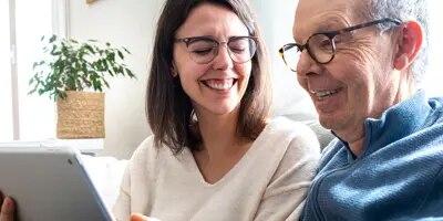 Une femme et un homme plus âgé rient en partageant une tablette sur le canapé.