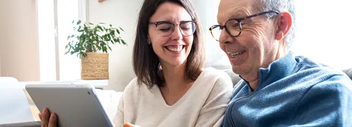 Une femme et un homme plus âgé rient en partageant une tablette sur le canapé.