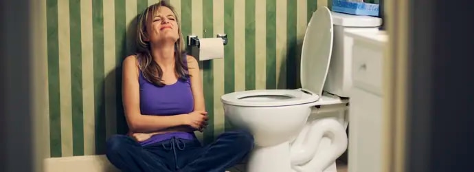 Femme assise par terre à côté des toilettes, se tenant le ventre de douleur