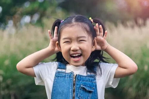 Une jeune fille sourit largement, au milieu d'un éclat de rire, en mettant ses mains derrière ses oreilles.
