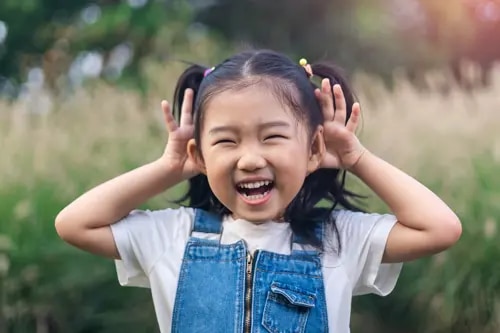 Une jeune fille sourit largement, au milieu d'un éclat de rire, en mettant ses mains derrière ses oreilles.