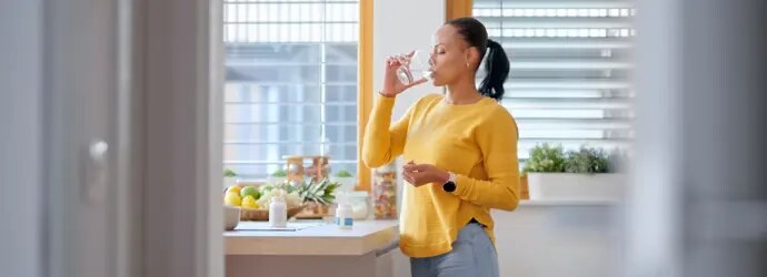 Une femme en pull jaune boit de l'eau dans la cuisine.