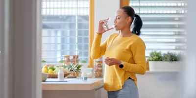 Une femme en pull jaune boit de l'eau dans la cuisine.