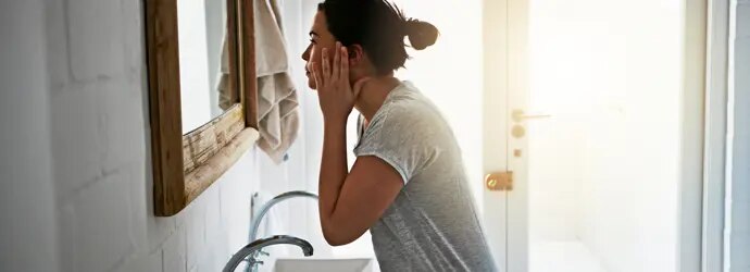 Une femme vêtue d'un haut gris se lave le visage au-dessus d'un lavabo blanc.