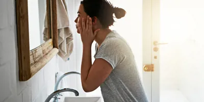 Une femme vêtue d'un haut gris se lave le visage au-dessus d'un lavabo blanc.