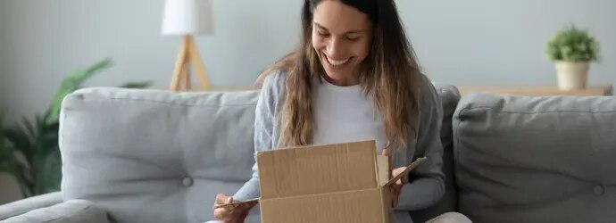 Une femme est sur un canapé gris, elle arbore un grand sourire en regardant à l’intérieur de son colis surprise.