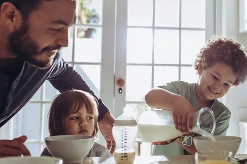 À la table du petit-déjeuner, un garçon verse du lait dans un bol, sous le regard de ses frères, sœurs et de son père.